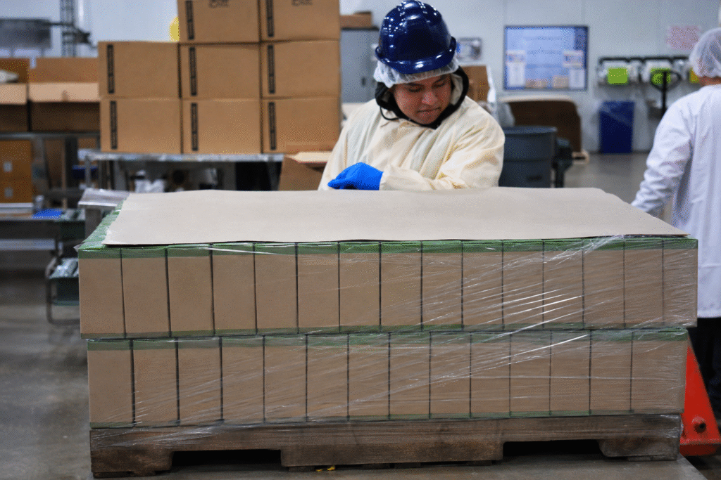 Worker inspecting a shrink-wrapped pallet of unbranded brown cardboard boxes in an APC production facility