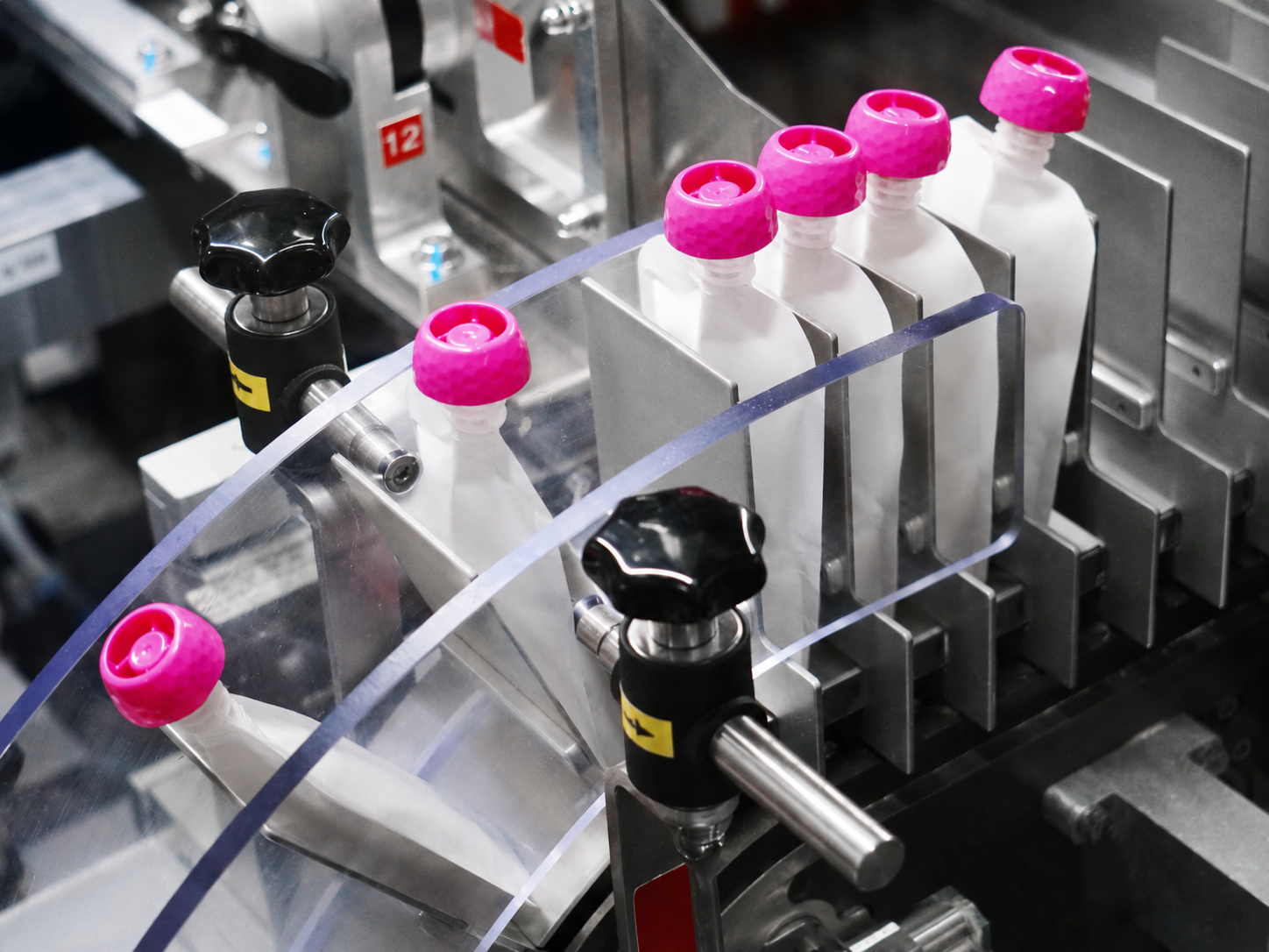 Unbranded white squeeze pouches with pink caps moving through automated packaging equipment at APC