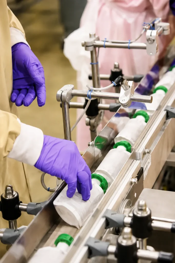 Worker wearing purple gloves handling unbranded white squeeze pouches with green caps on an automated APC packaging line