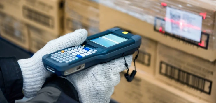 Gloved worker holding a handheld barcode scanner beside stacked cardboard cartons.