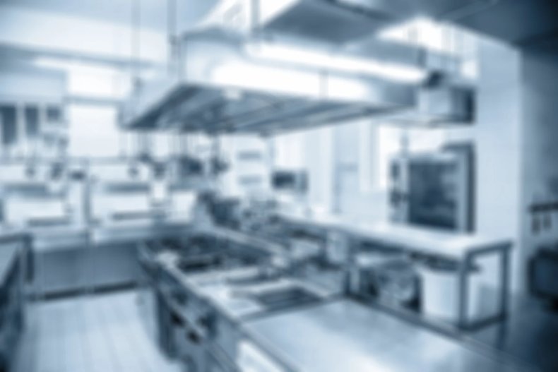Out-of-focus commercial kitchen with stainless steel counters, cooking stations, and an overhead exhaust hood, in a cool blue tone.