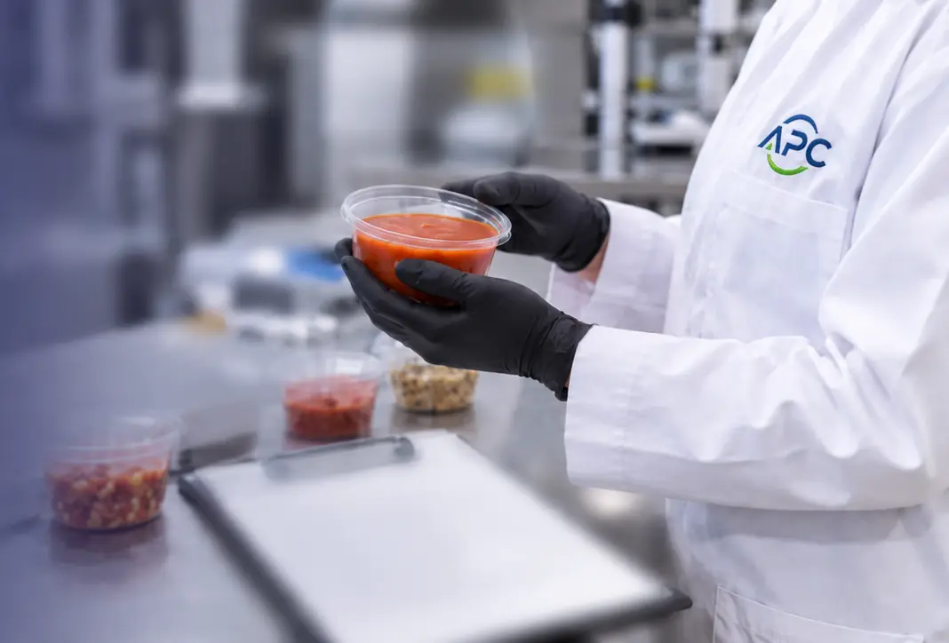 APC food safety specialist in a lab coat and black gloves holding a clear sealed cup of red sauce, with more sealed sample cups and a clipboard on a stainless-steel bench