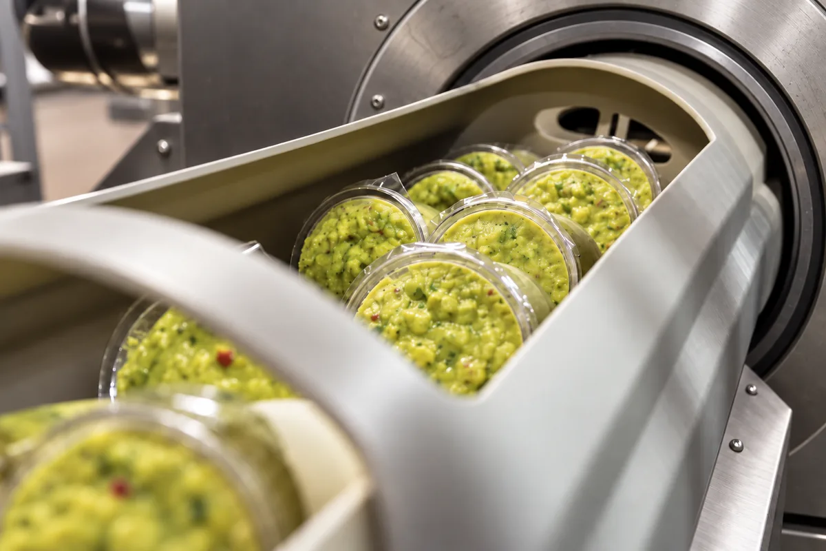 Clear cups of guacamole in a carrier entering a clean stainless steel HPP machine.