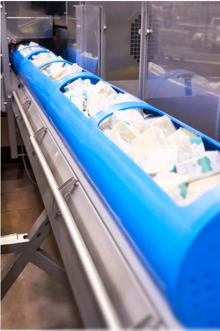 Sealed food packages loaded in blue perforated carriers on a stainless steel processing line